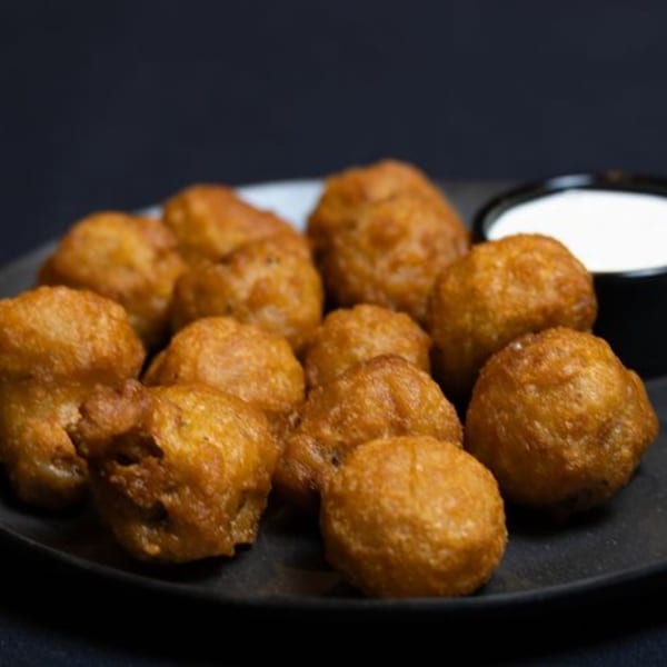 Golden-brown fried mushrooms are piled on a dark plate next to a small cup of white dipping sauce.