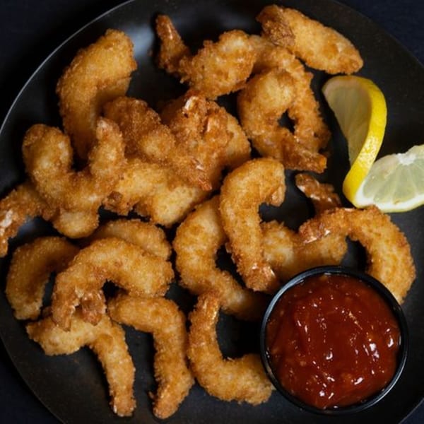 Crispy fried shrimp with two lemon wedges and a small bowl of red dipping sauce on a black plate.